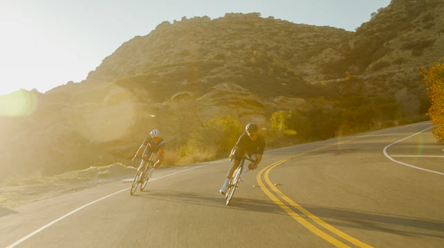 Dois ciclistas lado a lado numa estrada rural ao fim da tarde, contraluz dourado