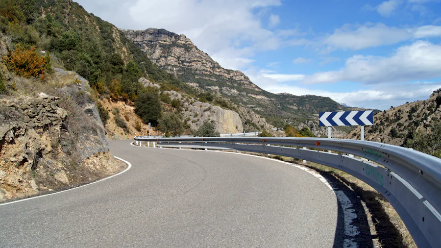 Estrada de montanha larga a contornar uma encosta, com barreiras de protecção e montanhas ao fundo